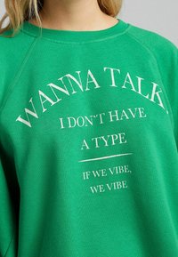 Zelená mikina se žebrovaným kulatým výstřihem. Obsahuje bílý text: "CHCEŠ MLUVIT, NEMÁM ŽÁDNÝ TYP, JESTLI SI SEDNEME, TAK SI SEDNEME." Měkký materiál.