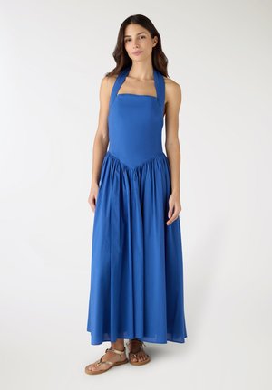 Donna in piedi che indossa un abito lungo blu senza maniche con collo alla coreana e sandali beige bassi, su uno sfondo bianco semplice.