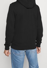 Černá mikina s kapucí z jemného materiálu, s žebrovanými manžetami a lemem. Nahoře je kombinovaná s světle modrými džínami, pohled zezadu.