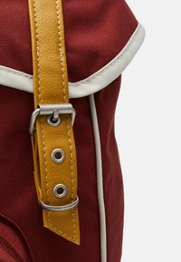 Sac en toile rouge avec une sangle en cuir jaune, des accessoires en métal argenté et des finitions blanches. La sangle est dotée d'une boucle et de boutons décoratifs.