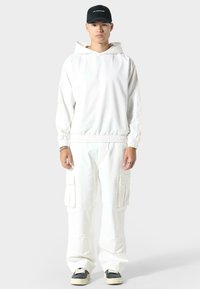 Felpa bianca con cappuccio e cintura elastica, abbinata a pantaloni cargo bianchi con tasche laterali. Cappellino nero con logo e scarpe nere.