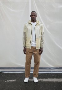 Translucent light yellow jacket with black zipper and logo, paired with tan trousers and white sneakers, set against a neutral backdrop.