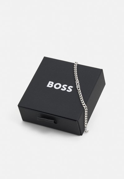 Bracelet en chaîne en argent posé sur une boîte noire portant le nom de la marque "BOSS" en lettres blanches en gras. La boîte dispose d'un onglet de traction pour l'ouvrir.