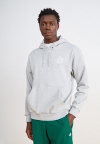 Halli hallide veste, ees taskuga ja nööriga kapuutsiga. Vasakul rinnal on väike valge logo. Mudel kannab rohelisi mustrilisi lühikesi pükse.