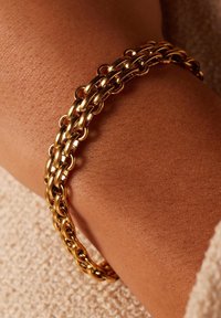 Gouden kettingarmband bestaande uit onderling verbonden ovale en ronde schakels, met een glanzende textuur, gedragen om een pols tegen een huidkleurige achtergrond.