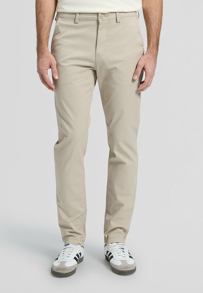 Pantalones de algodón beige con corte de pierna recta, cierre de botones y trabillas para cinturón, combinados con zapatillas blancas y negras.