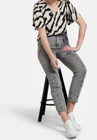 Top corto a maniche corte con stampa zebra bianca e nera, abbinato a jeans in denim grigio a vita alta e sneaker bianche, seduto su uno sgabello nero.