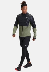 Chaqueta de zip de FILA con parte superior negra acolchada y parte inferior verde oliva, combinada con pantalones cortos negros y zapatos con acentos rojos.