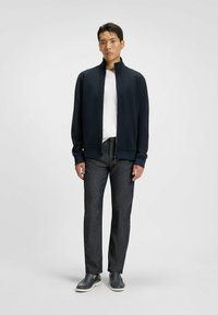 Marinblå zip-up jacka med hög krage, vit t-shirt under, mörka texturerade byxor och gråa slip-on skor. Ren, modern passform.