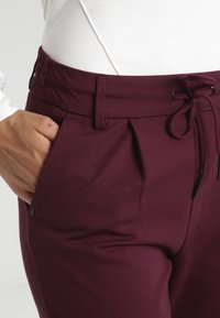 Mano en el bolsillo lateral de unos pantalones de tiro alto, color púrpura oscuro, con cordón y pasadores para cinturón, combinados con una blusa blanca.