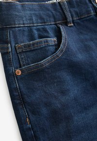 Donkerblauwe denim jeans met een voorzak met zichtbare stiksel en een koperen klinknagel detail, die een soepele textuur en een tailormade ontwerp benadrukken.