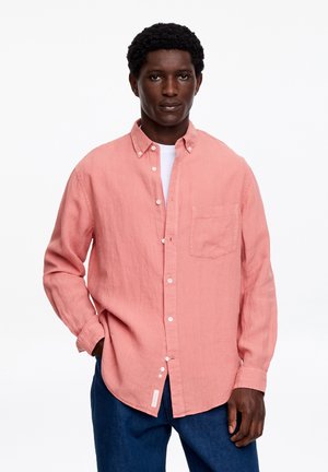 Mand iført en koralfarvet skjorte over en hvid T-shirt og mørkeblå bukser, stående med den ene hånd i lommen mod en hvid baggrund.