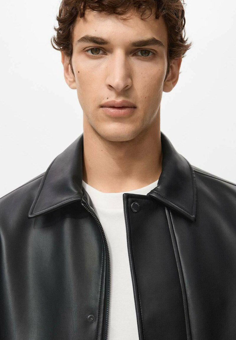 Schwarze Lederjacke mit glatter Textur, ausgestattet mit spitzem Kragen und Frontreißverschluss. Über einem weißen Hemd getragen.