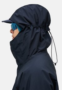 Giacca impermeabile blu navy con colletto alto e cappuccio regolabile con cordino. Presenta un cappello con visiera e dettagli con zip esterni.