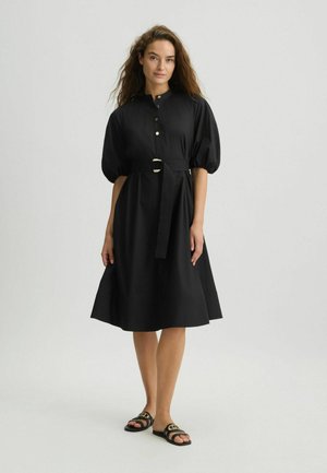 Een vrouw staat glimlachend in een zwarte jurk met opgezette mouwen en een riem, gecombineerd met platte sandalen, tegen een neutrale achtergrond.