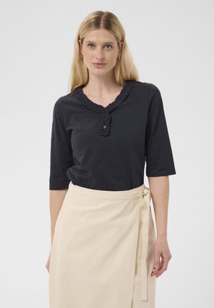 Femme aux cheveux blonds raides portant une chemise noire à col à volants et une jupe portefeuille beige nouée à la taille sur un fond uni.