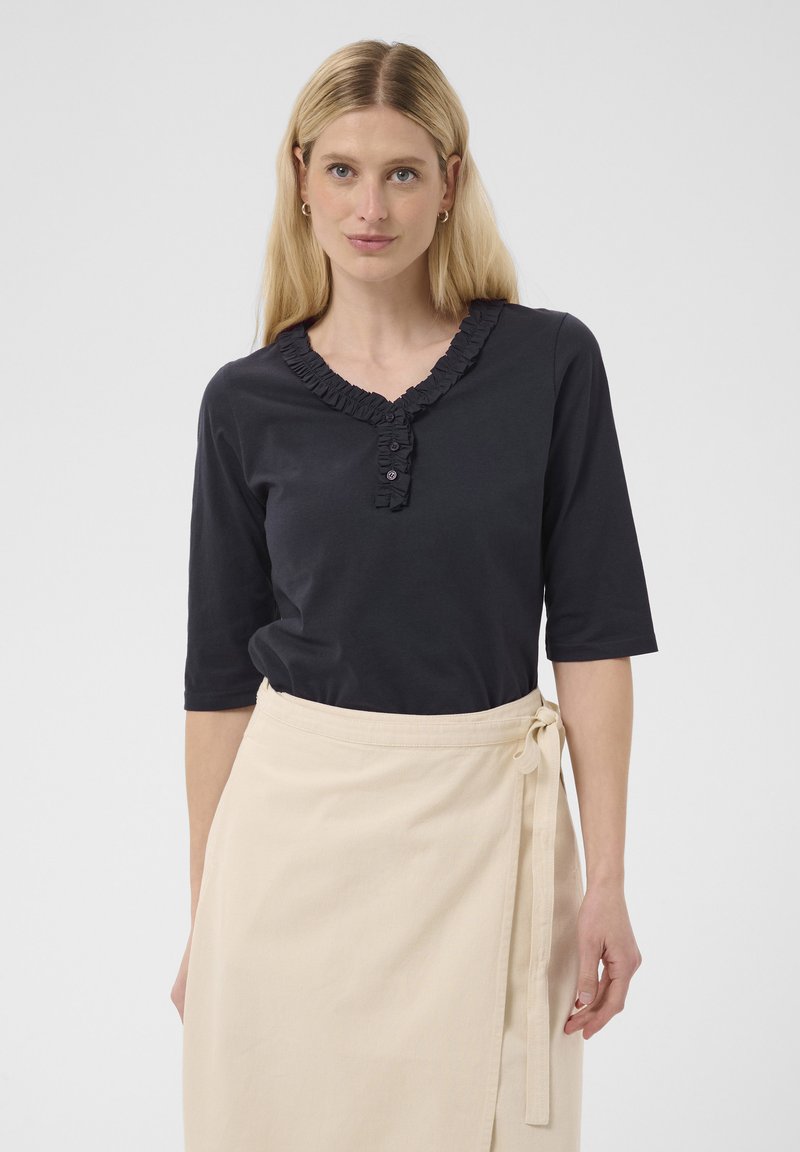 Femme aux cheveux blonds raides portant une chemise noire à col à volants et une jupe portefeuille beige nouée à la taille sur un fond uni.