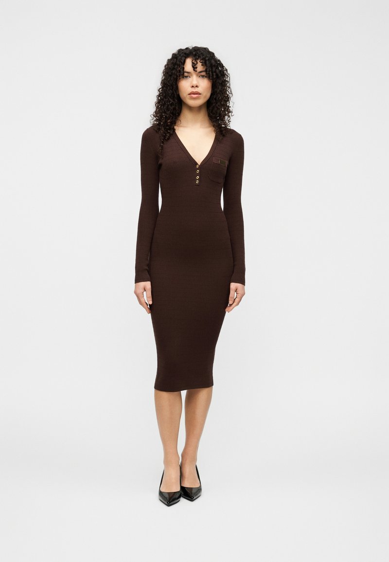 Καφέ, μακρυμάνικο φόρεμα με ριμπ ύφανση, V-λαιμόκοψη, κουμπιά και μήκος μέχρι το γόνατο. Φορεμένο με μαύρες ψηλοτάκουνες γόβες.