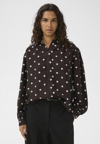 Rjava srajca z dolgimi rokavi, spredaj z gumbi in mehko teksturo. Ima bele pikice različnih velikosti za kontrast.