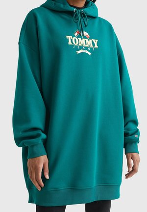 Personne portant un sweat à capuche ample de couleur bleu sarcelle avec le logo "Tommy Jeans", capuche à cordon et poignets ainsi qu'ourlet côtelés, sur un fond uni.