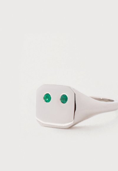Ezüst gyűrű lapos, téglalap alakú tetején két kis, kerek zöld drágakő egymás mellett, fehér háttéren.