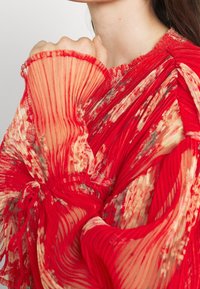 Punainen taitettu paita, joka on valmistettu läpinäkyvästä, tekstuurista kankaasta. Varustettu kukkakoristeilla, röyhelökauluksella ja suurilla hihoilla, joissa on elastiset hihansuut.