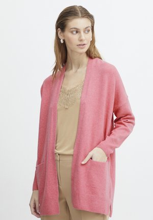 Cardigan rose en maille douce, à ouverture devant, avec des poches profondes et des poignets côtelés. Porté sur un top beige avec des bordures en dentelle.