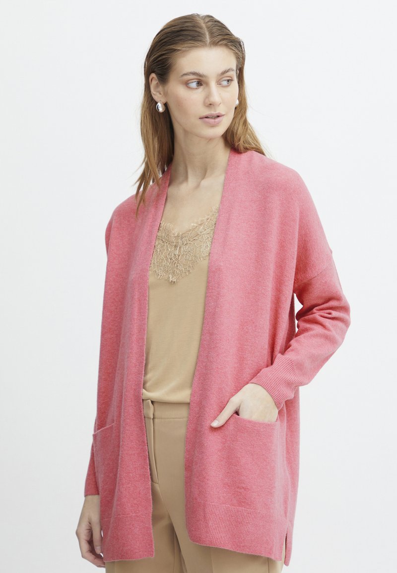 Cardigan rosa realizzato in morbido tessuto a maglia, con apertura frontale, profonde tasche e polsini a coste. Indossato sopra una maglietta beige con bordo in pizzo.