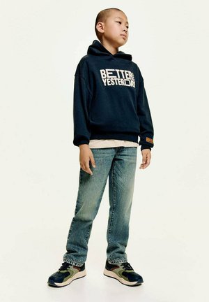 Felpa navy con testo "MEGLIO DI IERI", camicia chiara sotto, abbinata a jeans blu e sneakers multicolori. Pose in piedi.