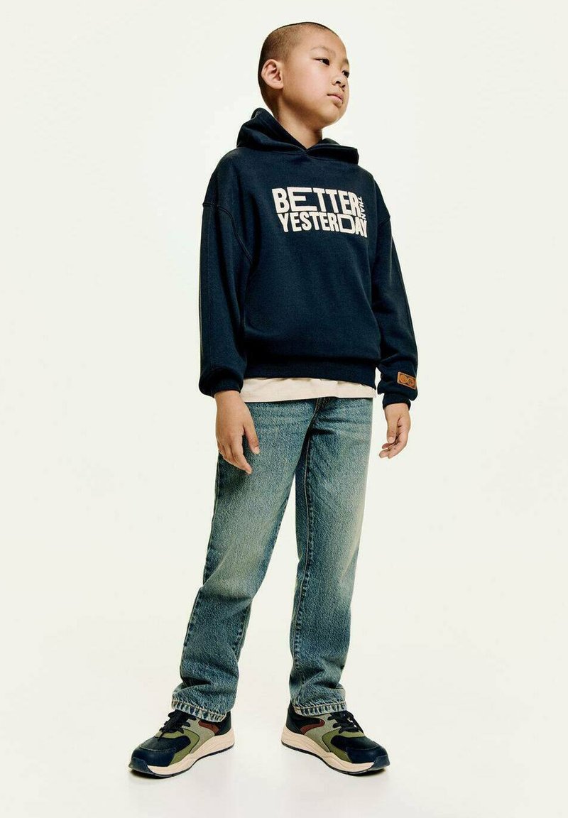 Marine hoodie met de tekst "BETER DAN GISTEREN", lichte shirt eronder, gecombineerd met blauwe jeans en multicolore sneakers. Staande pose.