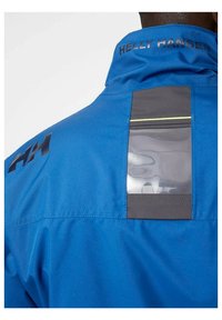 Blauwe waterdichte jas met hoge kraag, zwarte logo accenten en een transparante zak op de achterkant met een grijze rand en reflecterende details.