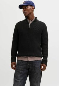 Schwarzer Strickpullover mit einem Viertel-Reißverschluss-Kragen, getragen über einem karierten Hemd, kombiniert mit dunklen Jeans. Legerer Lagenlook.