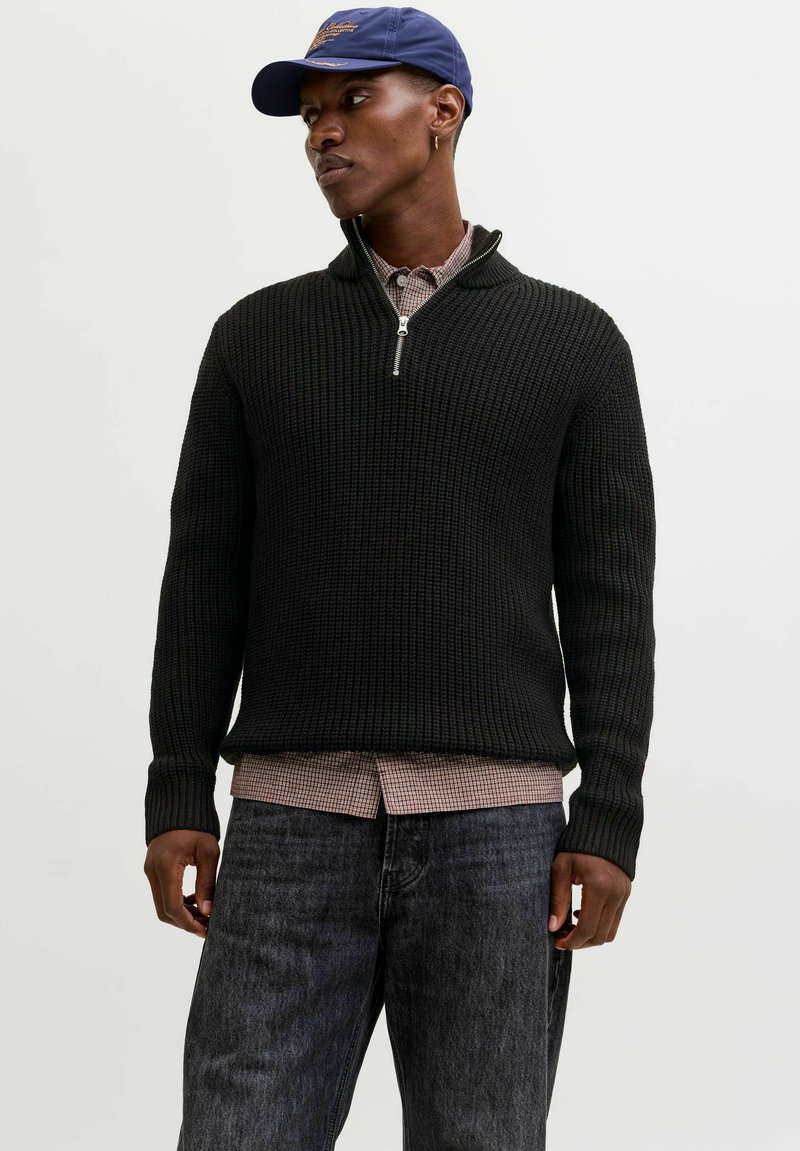 Schwarzer Strickpullover mit einem Viertel-Reißverschluss-Kragen, getragen über einem karierten Hemd, kombiniert mit dunklen Jeans. Legerer Lagenlook.