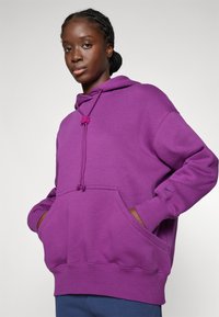 Lilla hettegenser med lommes front, snøring og ribbestrikkede mansjetter. Har en liten logo på ermet. Myk stofftekstur.