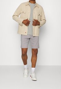 Beige canvasjakke med to brystlommer, stribet grå og hvid t-shirt, lysegrå shorts og hvide sneakers med røde detaljer.