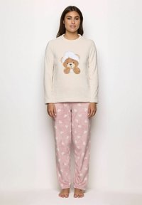 Una donna che indossa un top a maniche lunghe color crema con una grafica di orsetto di peluche e pantaloni da pigiama in pile rosa con motivi a cuori bianchi, in piedi a piedi nudi.
