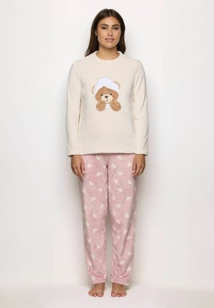 Mujer vistiendo una blusa de mangas largas color crema con un gráfico de oso de peluche y pantalones de pijama de forro polar rosa con patrones de corazones blancos, de pie descalza.