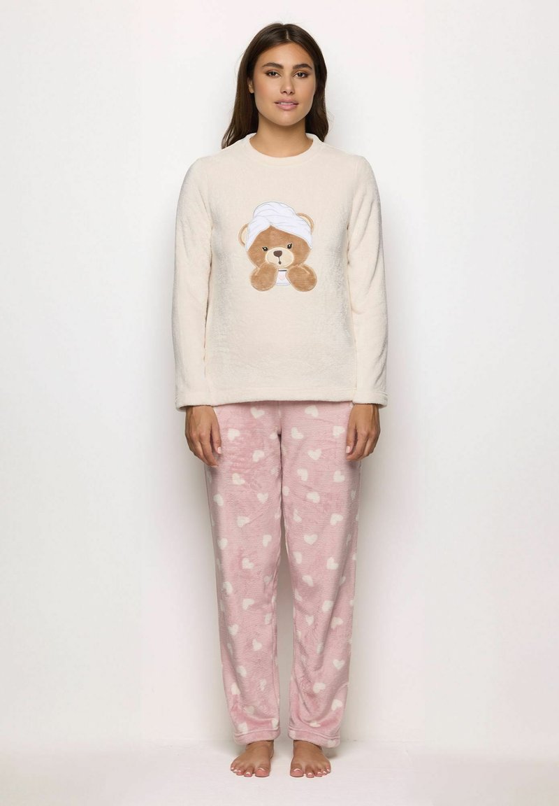 Una donna che indossa un top a maniche lunghe color crema con una grafica di orsetto di peluche e pantaloni da pigiama in pile rosa con motivi a cuori bianchi, in piedi a piedi nudi.