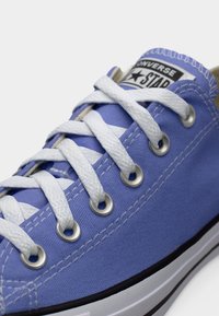 Κοντινή λήψη ενός ανοιχτόμωβ αθλητικού παπουτσιού Converse με λευκά κορδόνια και μεταλλικά κρικάκια, σε λευκό φόντο.