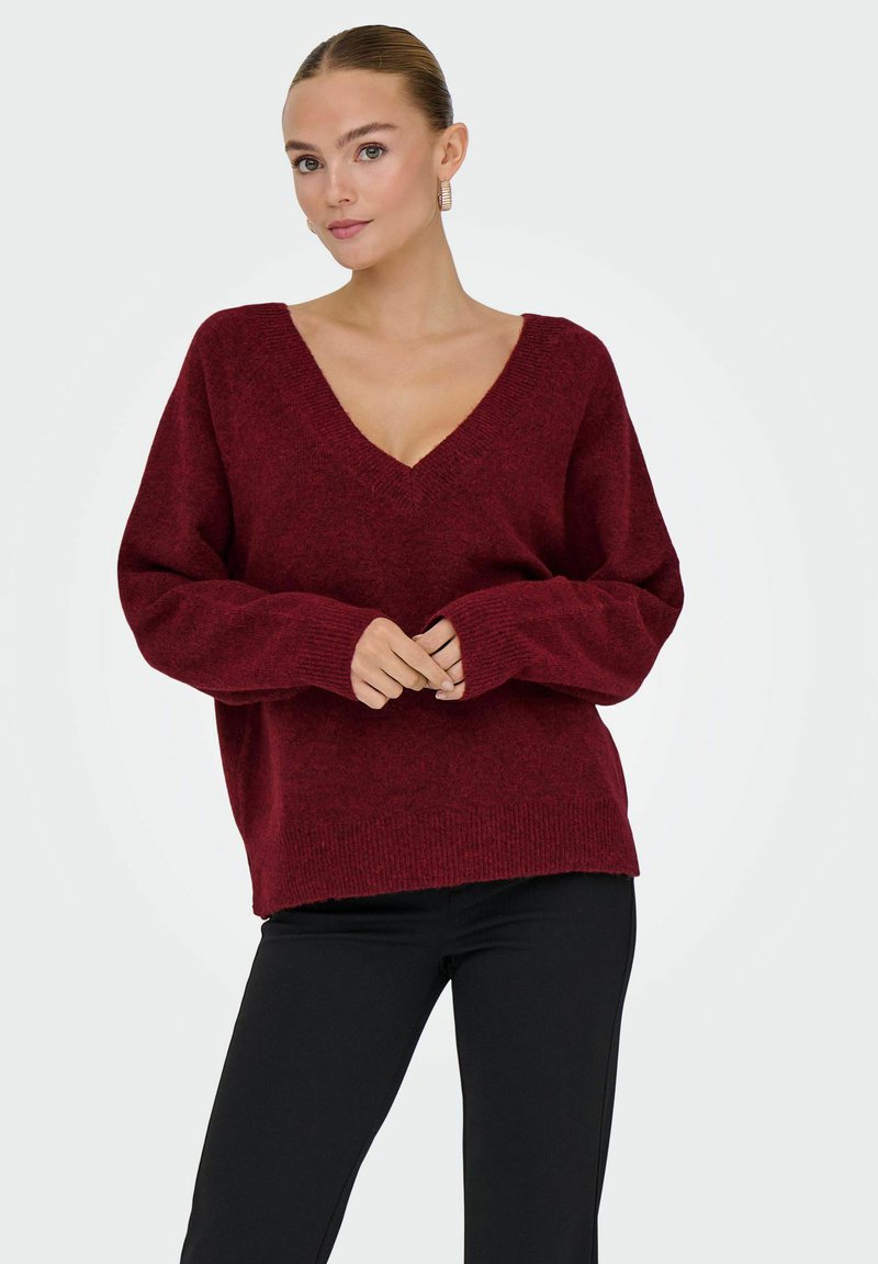 Pull en V bordeaux avec une coupe décontractée, manches longues, bord-côtes aux poignets et à l'ourlet, fabriqué en maille texturée. Associé à un pantalon noir.