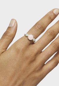 Anillo de cuarzo rosa con una piedra central redonda y facetada, flanqueada por acentos azul claro, montado en una sencilla banda de plata. Mano texturizada visible.