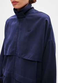 Giubbotto leggero di colore blu navy con colletto alto, caratterizzato da un design a strati e un logo ricamato sul petto. Texture liscia.