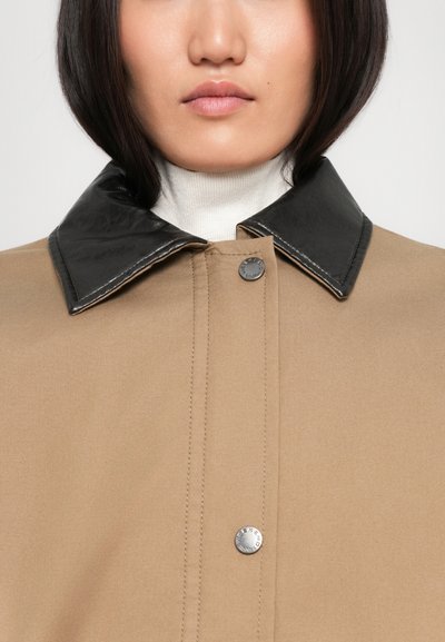 Manteau beige avec un col en cuir noir, présentant des boutons pressions en argent et une texture lisse. Design minimaliste sans motifs ni accents.