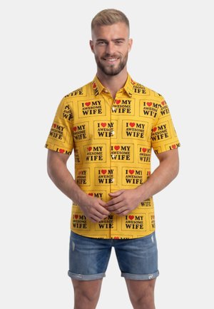 Uśmiechnięty mężczyzna z brodą ma na sobie żółtą koszulkę z nadrukiem "I love my awesome wife" i niebieskie dżinsowe szorty, stojąc na białym tle.