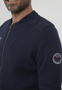 Tamsiai mėlynas megztinis su užtrauktuku, turintis gilią apykaklę ir rankogalius, bei ant kairiojo rankovicės esantis lopas su detaliu tekstu ir logotipu.