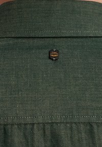 Col en chemise en tissu green herringbone avec une épingle de logo PME Legend noir et or située au centre du dos, présentant un motif texturé.