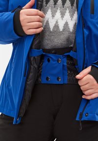 Chaqueta impermeable azul con acentos negros, que presenta un cierre de botón a presión, cinturilla elástica y un suéter gris de patrón debajo.