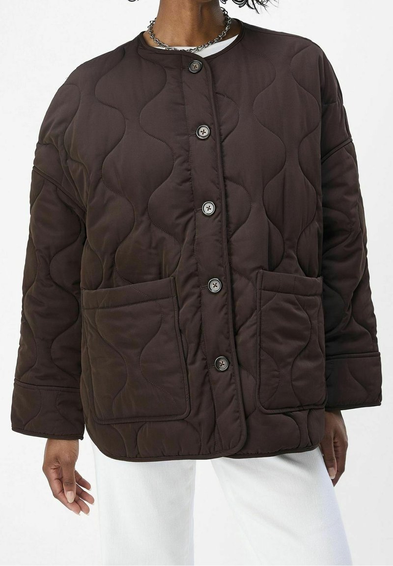 Personne portant une veste matelassée marron foncé avec des boutons visibles, de grandes poches avant et un pantalon blanc, vue du cou aux cuisses.