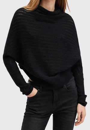 Femme portant un pull en maille noire ample avec des épaules tombantes et un jean noir ajusté, avec une main dans la poche sur un fond uni.