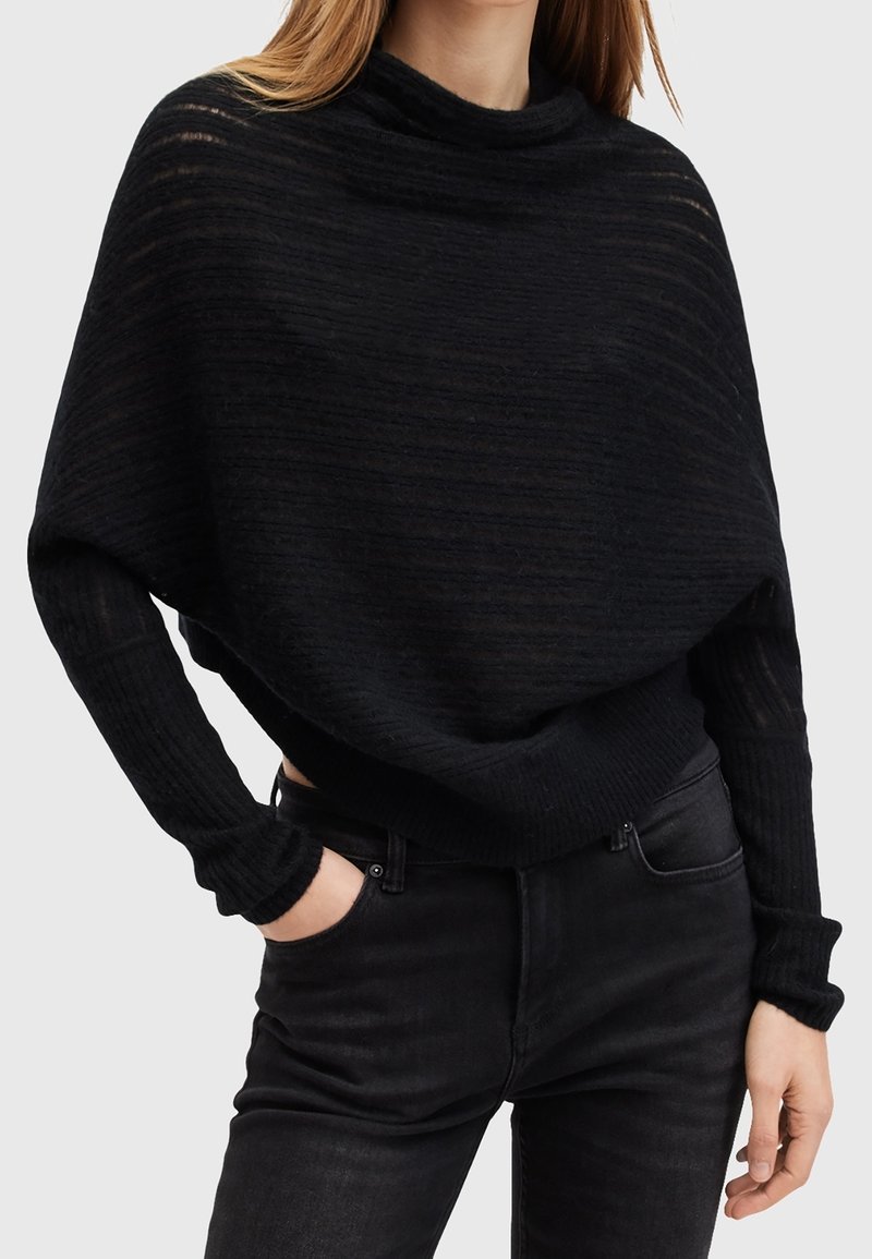 Femme portant un pull en maille noire ample avec des épaules tombantes et un jean noir ajusté, avec une main dans la poche sur un fond uni.